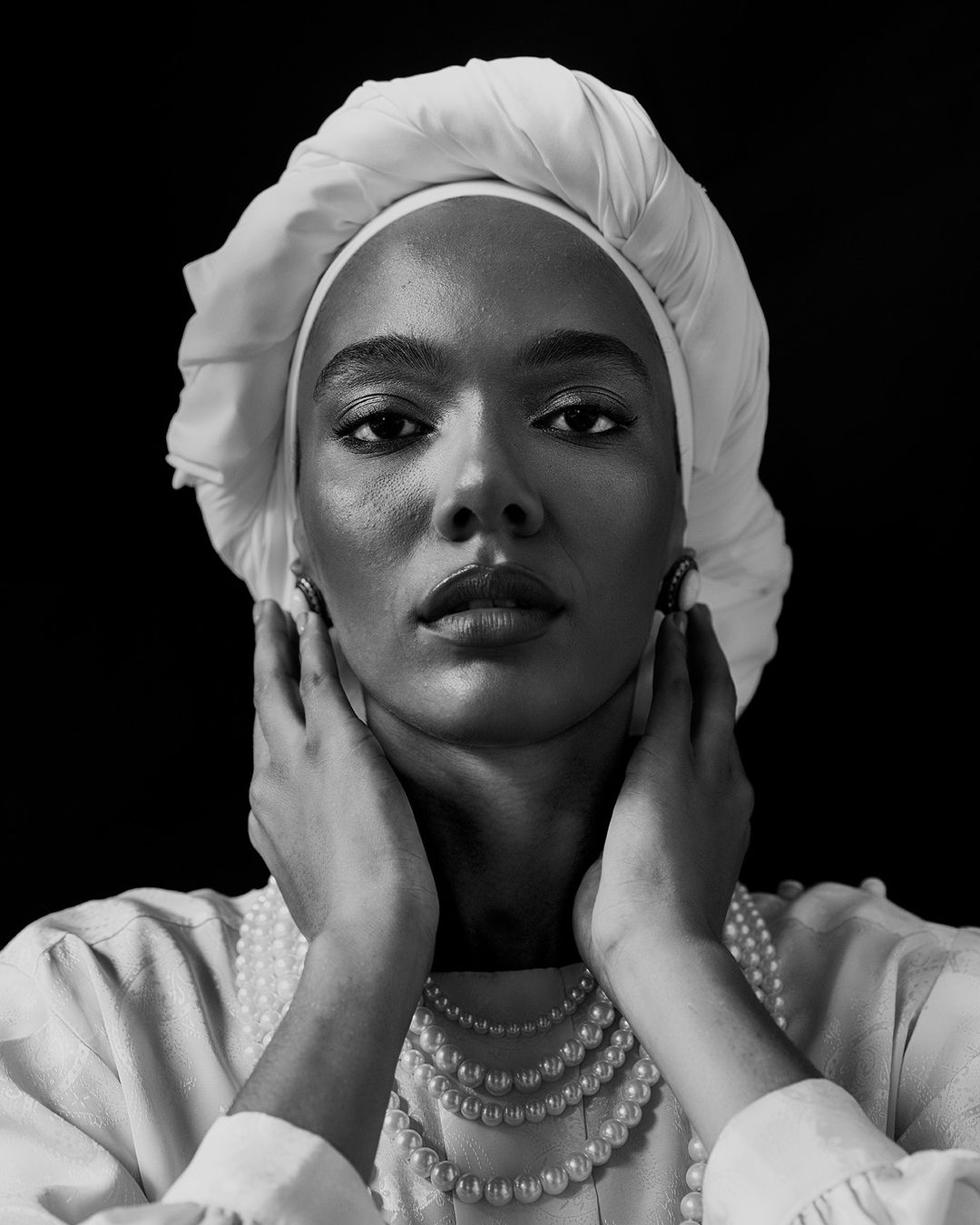 Black woman wearing a white turban and layered pearl necklaces touching her face in a monochrome portrait.