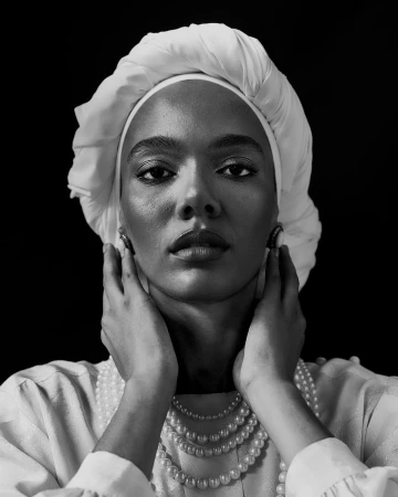 Black woman wearing a white turban and layered pearl necklaces touching her face in a monochrome portrait.
