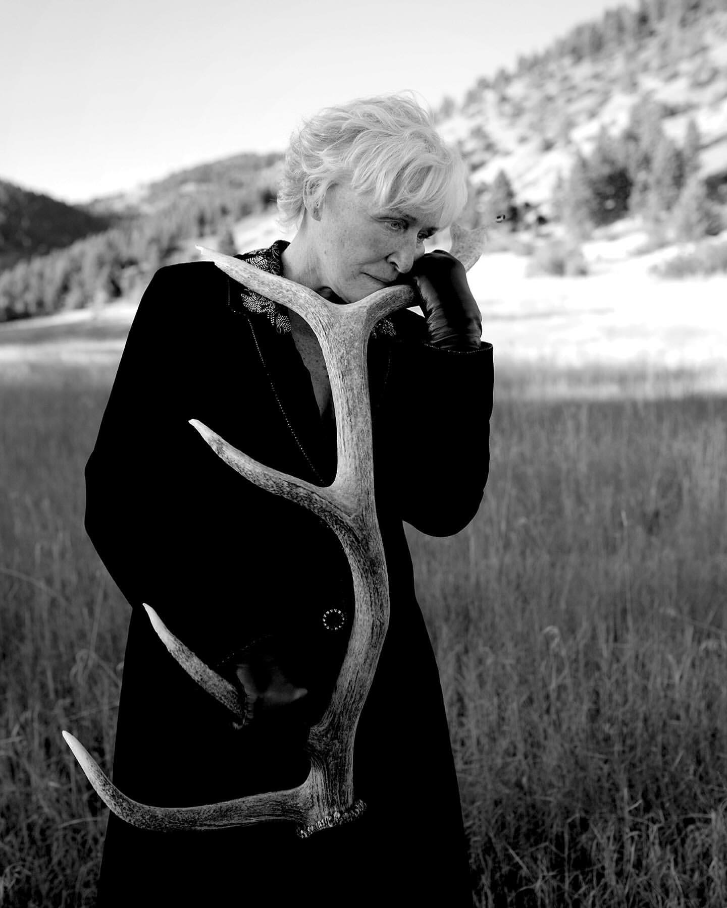 Black and white portrait of a mature woman outdoors wearing a dark coat, holding a large antler against a blurred mountain backdrop.