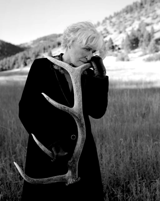Black and white portrait of a mature woman outdoors wearing a dark coat, holding a large antler against a blurred mountain backdrop.