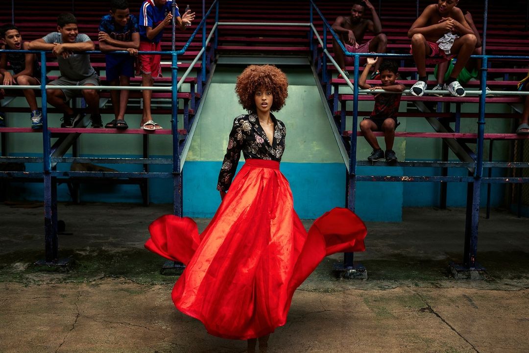 Woman in black floral top and flowing red skirt poses at an outdoor arena, surrounded by young men sitting on stadium-style seating.
