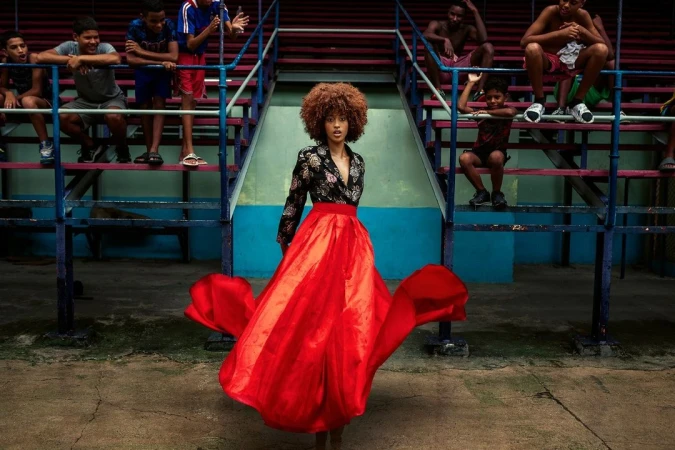 Woman in black floral top and flowing red skirt poses at an outdoor arena, surrounded by young men sitting on stadium-style seating.