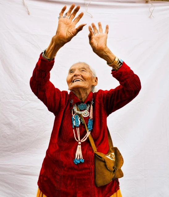 Elderly Indigenous woman in red shirt smiles with arms raised against a white backdrop, adorned with turquoise jewelry and a brown bag.