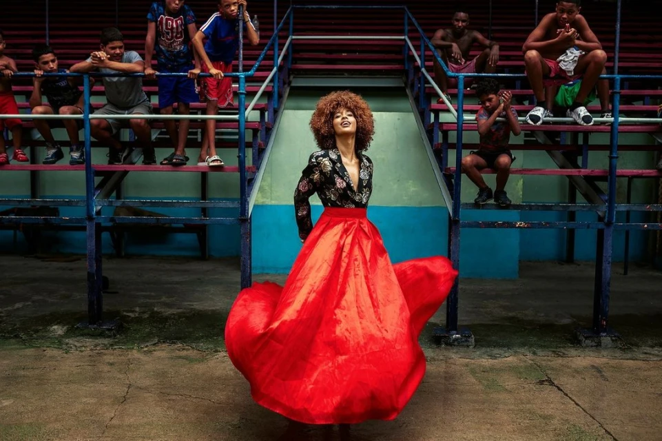 Fashion shoot featuring a Black woman in a vibrant red gown and floral blazer, posing outdoors in front of an audience on stadium bleachers.