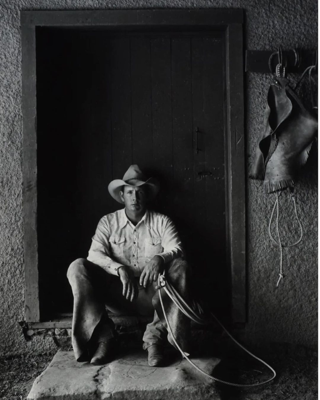 Monochromatic portrait of a cowboy sitting in a doorway, wearing a cowboy hat and holding a rope, with leather chaps hanging on the wall.