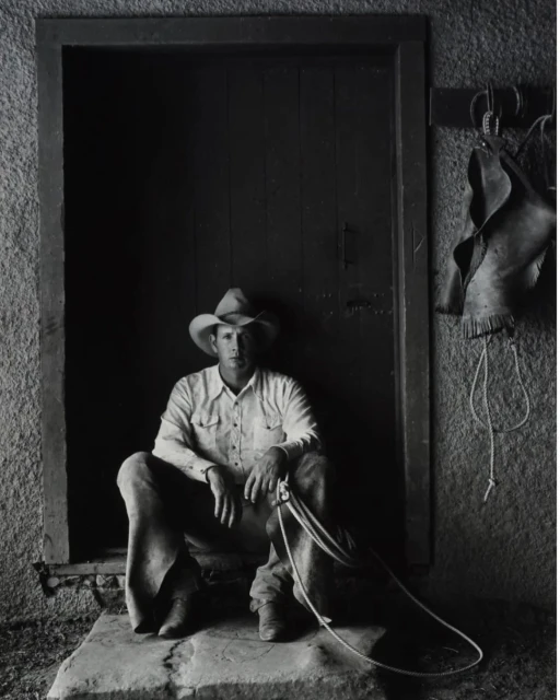 Monochromatic portrait of a cowboy sitting in a doorway, wearing a cowboy hat and holding a rope, with leather chaps hanging on the wall.