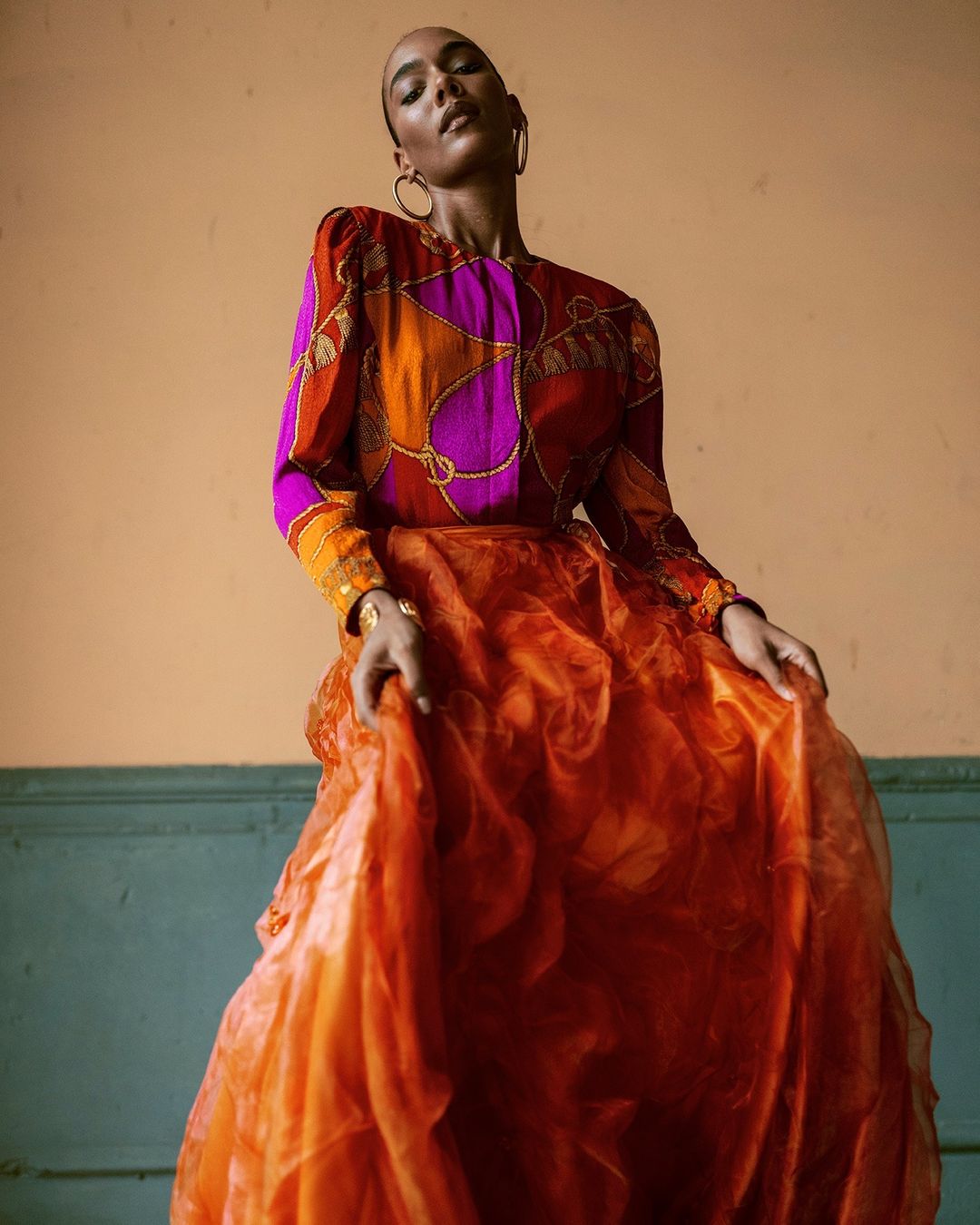Model in printed magenta and orange silk blouse and voluminous orange skirt posing against a painted interior wall.
