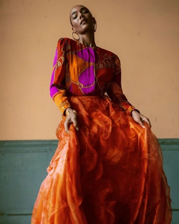 Model in printed magenta and orange silk blouse and voluminous orange skirt posing against a painted interior wall.