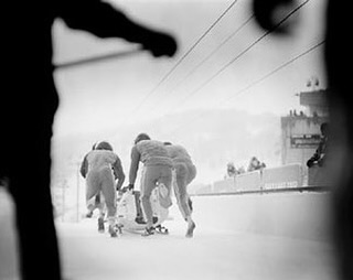 Vintage black and white image of a group of people pushing a bobsled on an icy track, with mountains in the background and overhead cables.