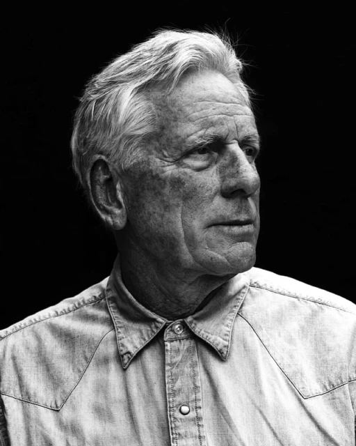 Monochrome portrait of a mature adult man with gray hair wearing a collared shirt against a dark background, looking off to the side.