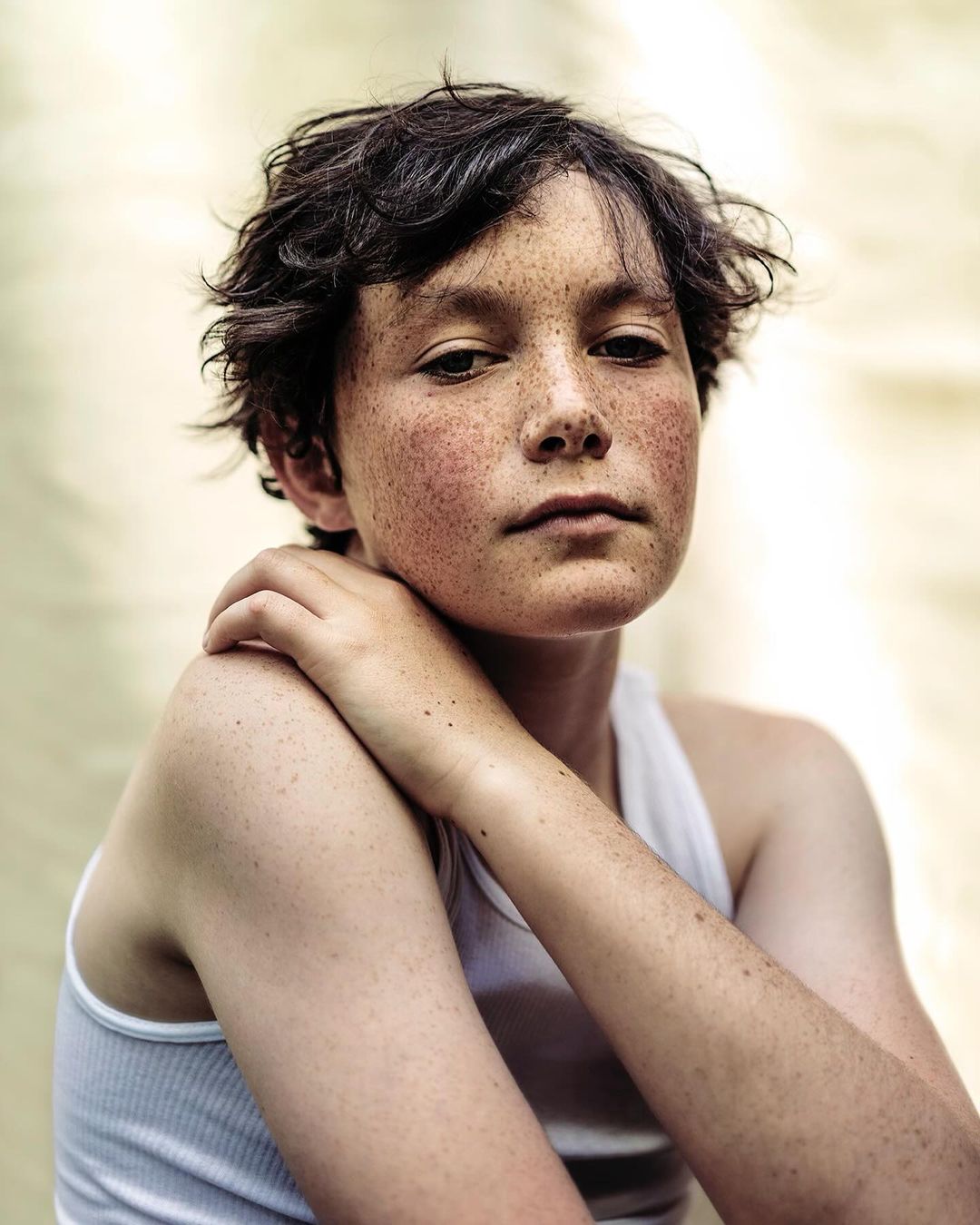 Portrait of a young freckled person in a white tank top, looking directly at the camera with hand on shoulder, against a neutral backdrop.