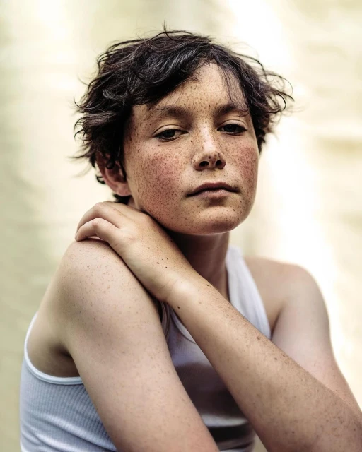 Portrait of a young freckled person in a white tank top, looking directly at the camera with hand on shoulder, against a neutral backdrop.