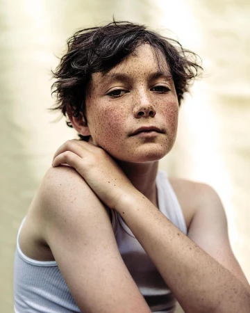 Portrait of a young freckled person in a white tank top, looking directly at the camera with hand on shoulder, against a neutral backdrop.
