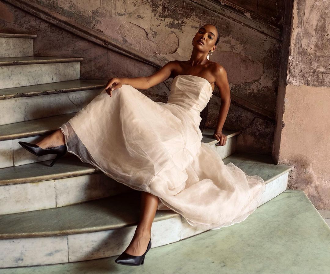 Elegant Black woman in a formal gown poses on marble stairs with black heels in an indoor setting, showcasing fashion and style.