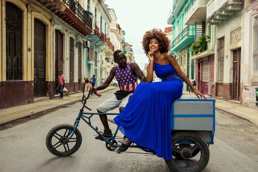 Two people on a bicycle rickshaw traveling down a city street with colorful buildings in Havana, Cuba on a sunny day. 