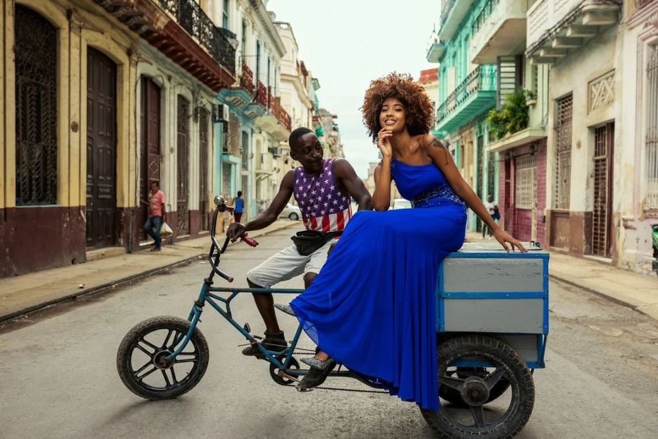 Two people on a bicycle rickshaw traveling down a city street with colorful buildings in Havana, Cuba on a sunny day. 