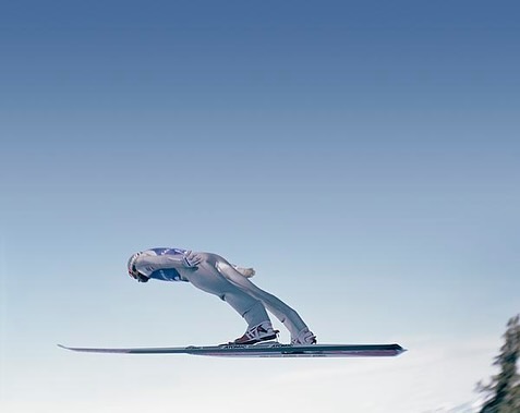 Action shot of an adult male ski jumper in mid-air, skis extended, against a clear blue sky with a hint of snow-covered ground.