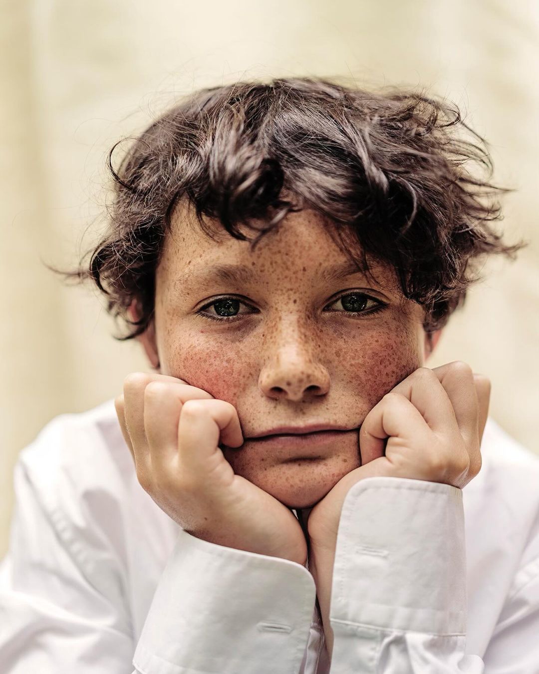 Portrait of a young person with freckles, looking at the camera with hands under their chin, wearing a white collared shirt.