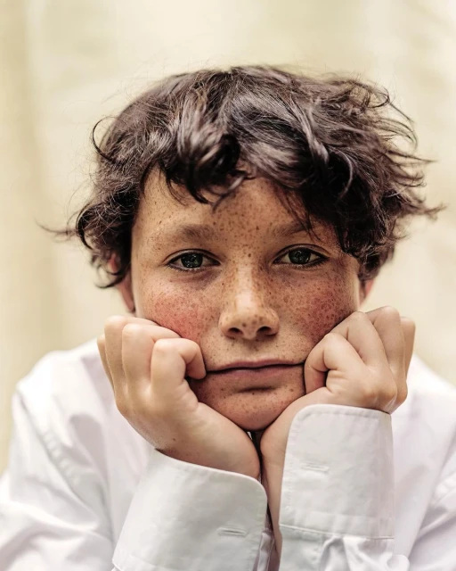 Portrait of a young person with freckles, looking at the camera with hands under their chin, wearing a white collared shirt.