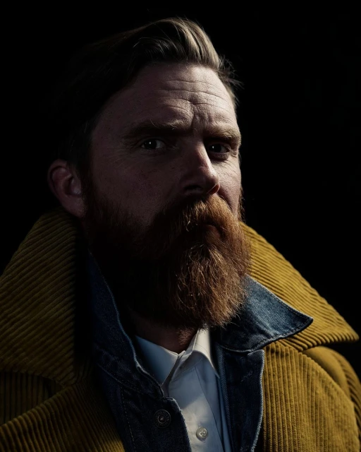 Portrait of a man with a red beard wearing a mustard-colored corduroy jacket and denim collar against a dark, shadowed background.