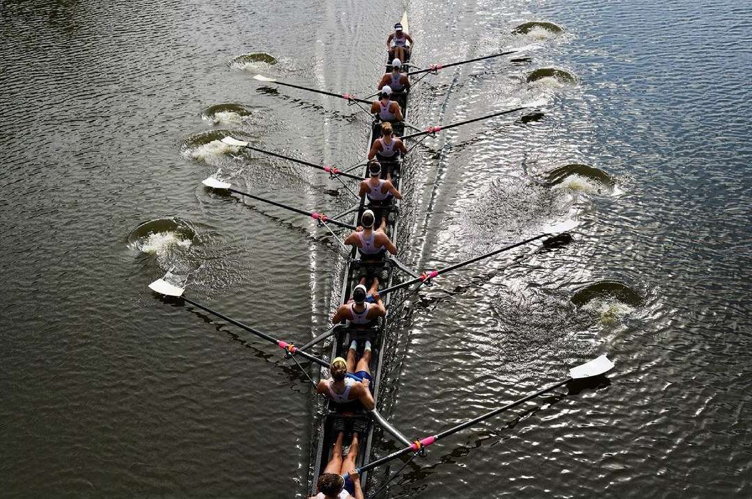 Overhead aerial view of eight athletes in a sculling boat on a dark river, propelling themselves with long oars creating splashes.