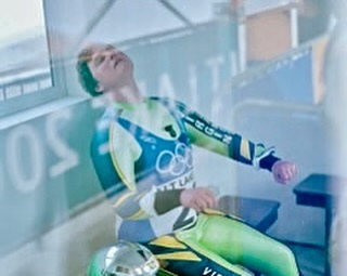 Athlete in green and yellow uniform sits exhausted in indoor setting, looking upward with a helmet beside them, visible through a window.