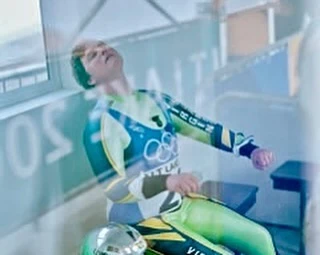 Athlete in green and yellow uniform sits exhausted in indoor setting, looking upward with a helmet beside them, visible through a window.