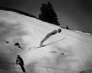 Black and white image of a ski jumper in mid-air against a snowy mountain backdrop, showcasing winter sports and athleticism.