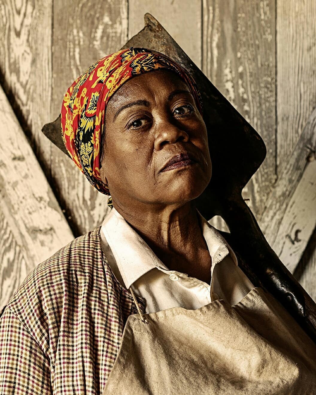 Portrait of an adult African-American woman in apron and headscarf, looking toward the camera, with tool over shoulder, against wooden background.