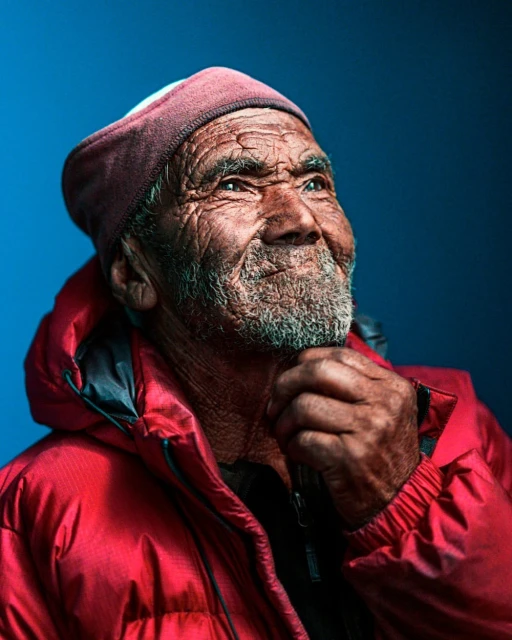 Portrait of an elderly man with weathered skin and a beanie, wearing a red puffer jacket, looking upwards against a blue background.
