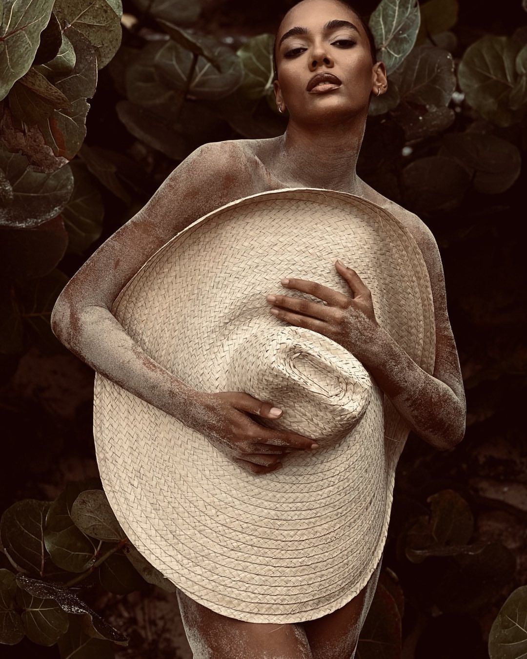 Portrait of a woman partially covered in sand, holding a large sun hat against a leafy background in an outdoor setting.