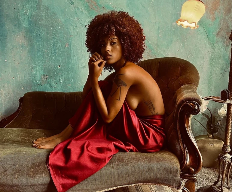 Artistic portrait of a nude Black woman draped in red fabric, seated on vintage green sofa against a textured turquoise wall with soft light.