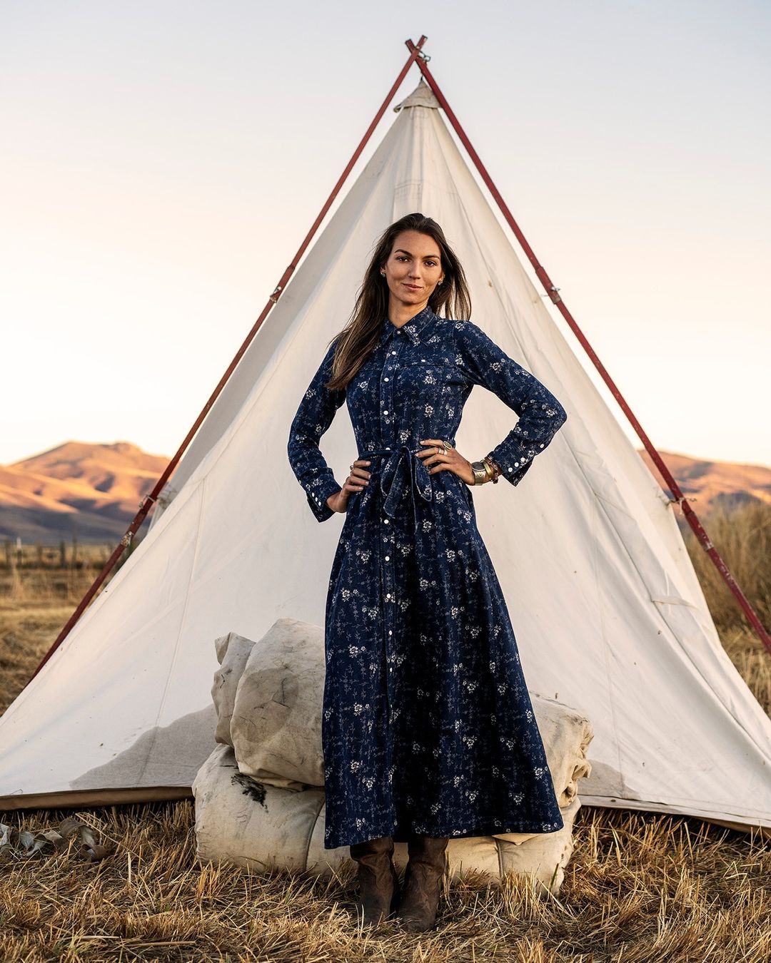 Adult female posing in long-sleeve floral maxi dress with hands on hips and white tent outdoors in a rural environment at sunset.