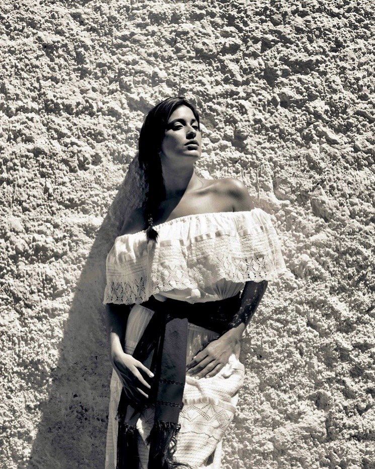 Black and white portrait of a woman in an off-the-shoulder dress leaning against a textured wall in natural lighting with dark hair.