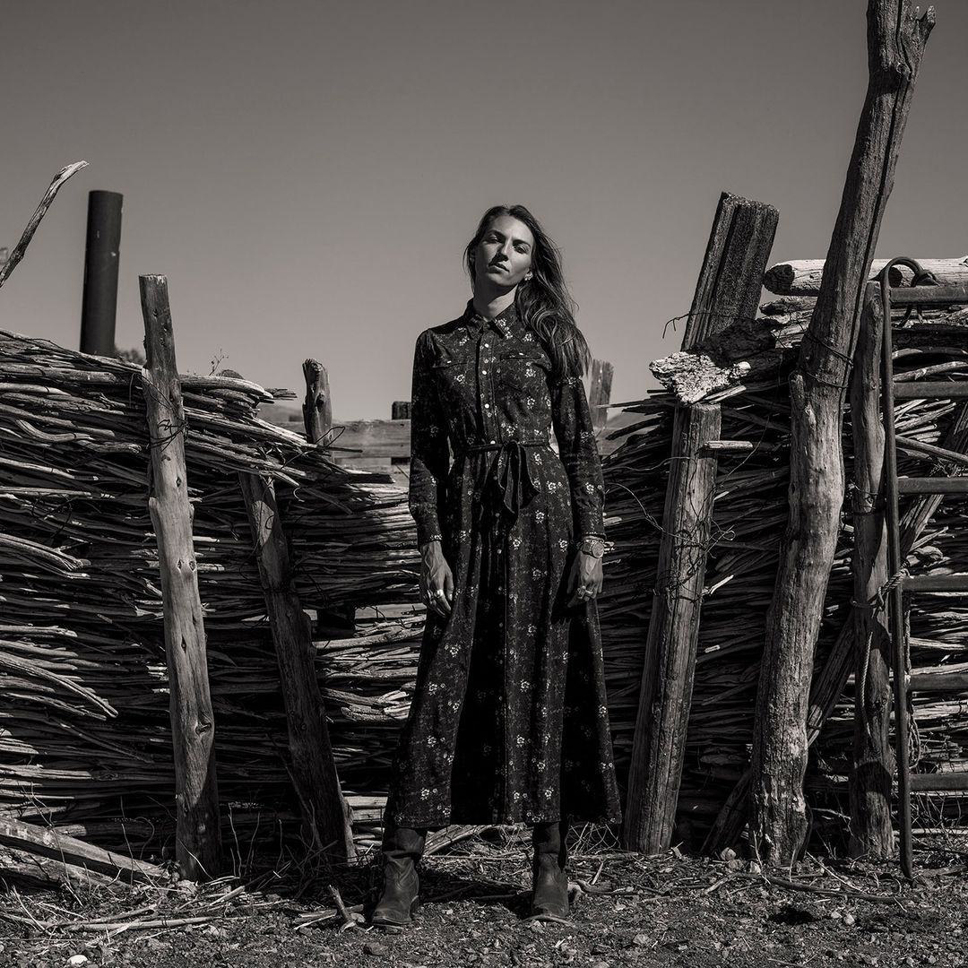 Black and white fashion shot of a woman in a long floral dress standing in front of a rustic wooden fence in a dry, outdoor landscape.