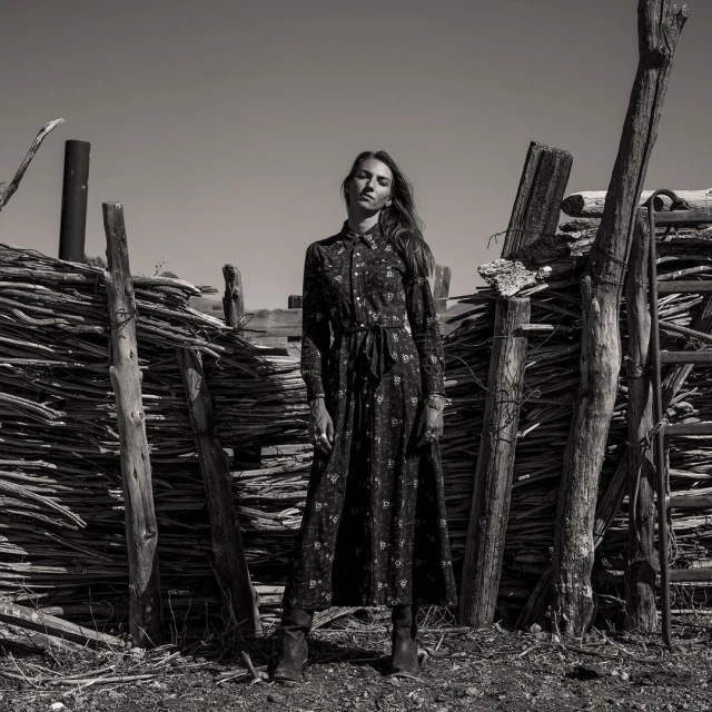 Black and white fashion shot of a woman in a long floral dress standing in front of a rustic wooden fence in a dry, outdoor landscape.