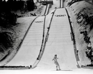 Monochrome shot of a skier on a ski jump ramp featuring the Olympic rings logo, snow-covered mountains, and a crowd in the background.