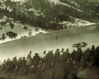 Vintage photo of a male luger racing down an icy track at the Winter Olympics, surrounded by a crowd in a snowy, mountainous landscape.