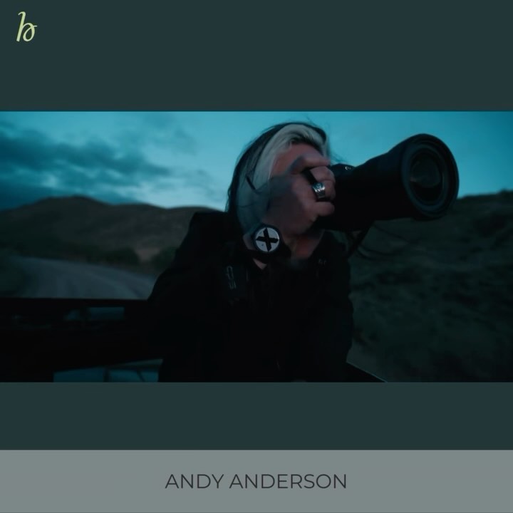 Photographer with a large telephoto lens on a rural road under cloudy skies. He is wearing a black jacket and has black and white hair.