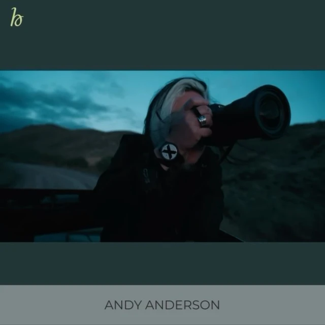 Photographer with a large telephoto lens on a rural road under cloudy skies. He is wearing a black jacket and has black and white hair.