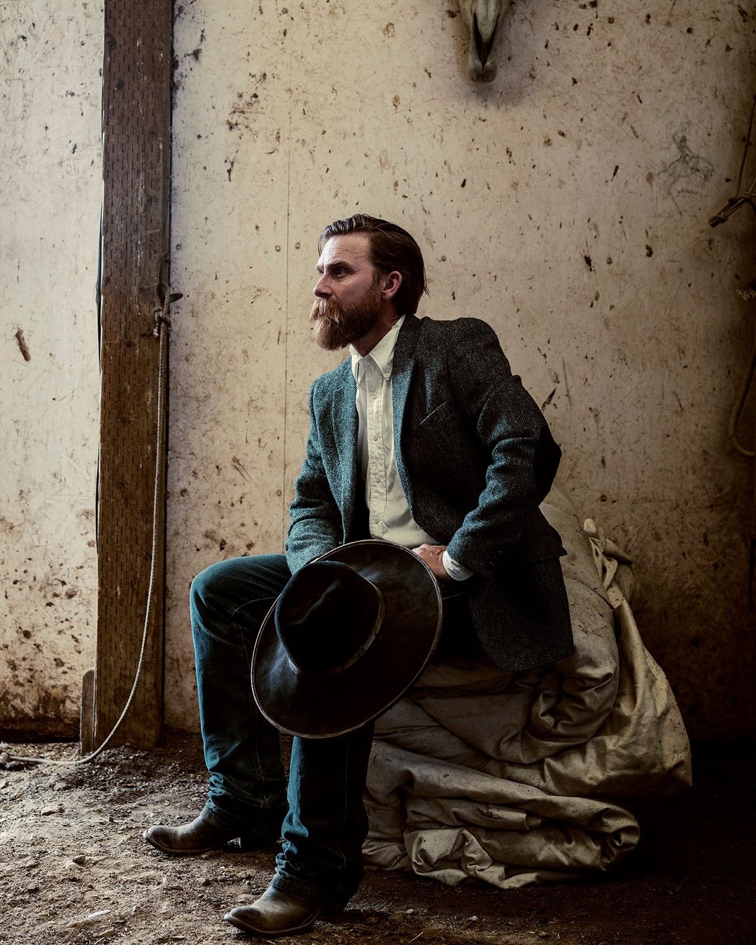 Bearded man in jeans and blazer sits with cowboy hat in barn, creating a vintage Western aesthetic with weathered wood and animal skull.