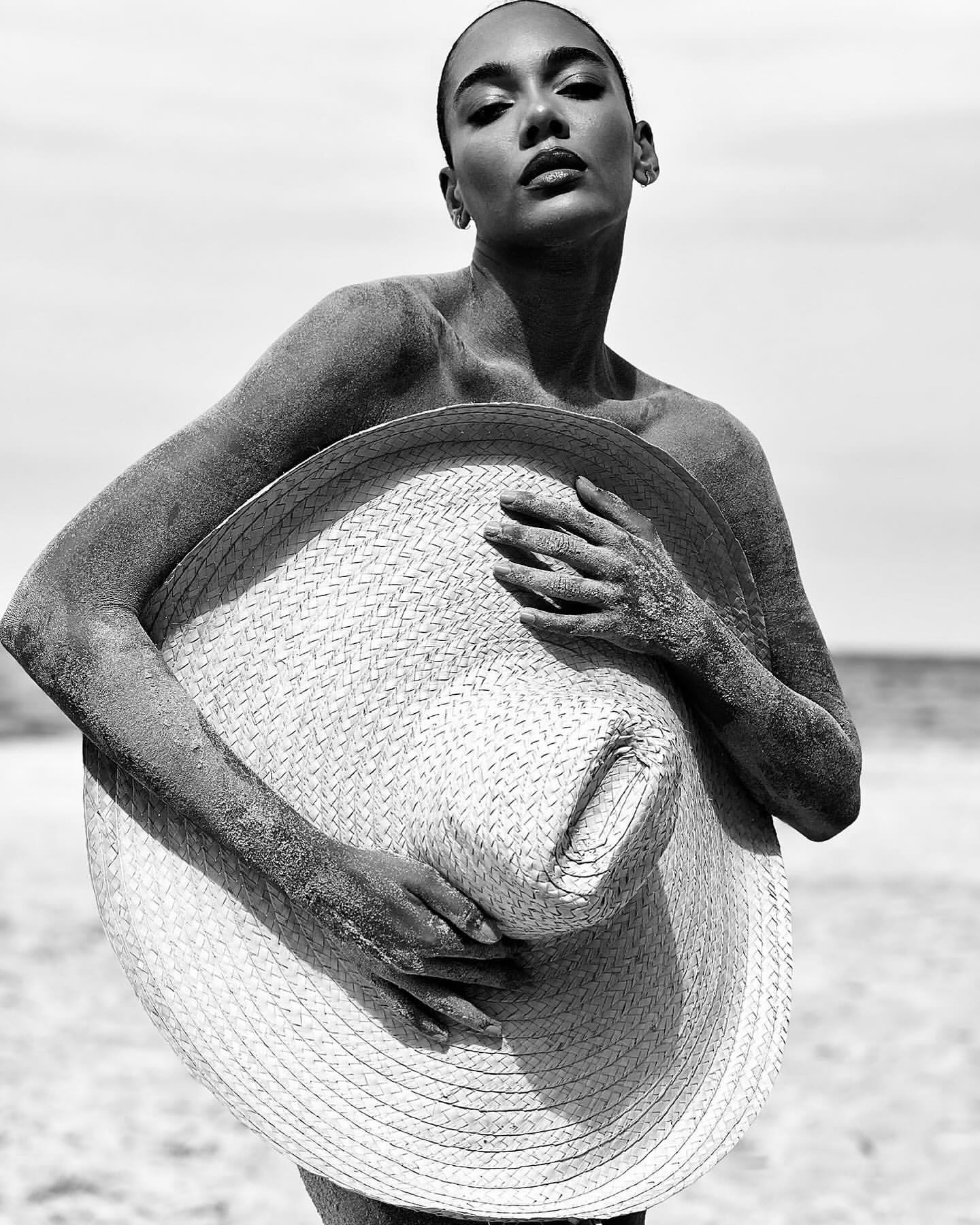 Black and white portrait of a woman on a beach, covered in sand, holding a straw hat against a sky and ocean background.