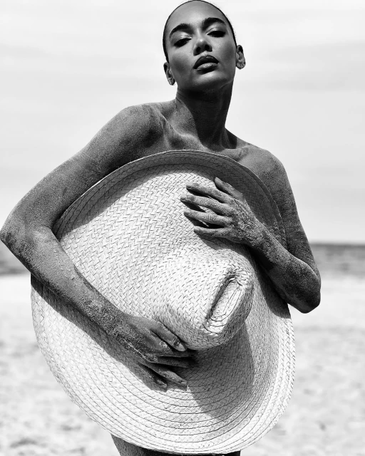 Black and white portrait of a woman on a beach, covered in sand, holding a straw hat against a sky and ocean background.
