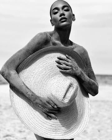 Black and white portrait of a woman on a beach, covered in sand, holding a straw hat against a sky and ocean background.
