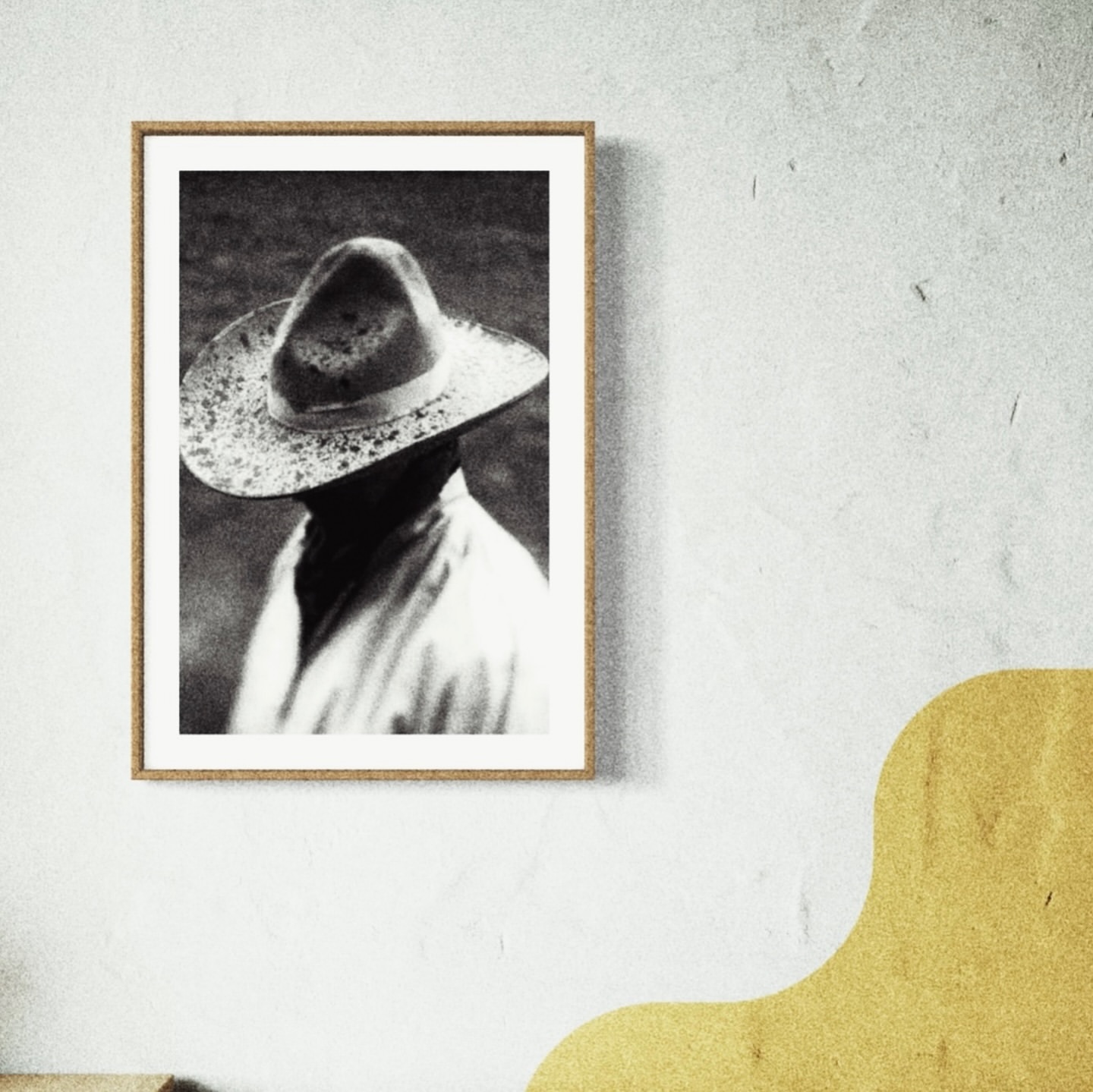Black and white photograph of a person wearing a hat, displayed in a picture frame on a textured white wall with yellow graphic.
