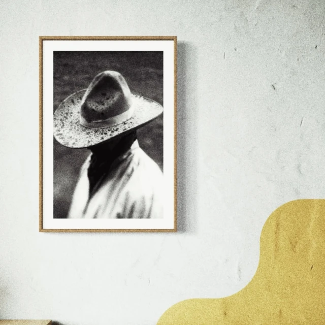 Black and white photograph of a person wearing a hat, displayed in a picture frame on a textured white wall with yellow graphic.