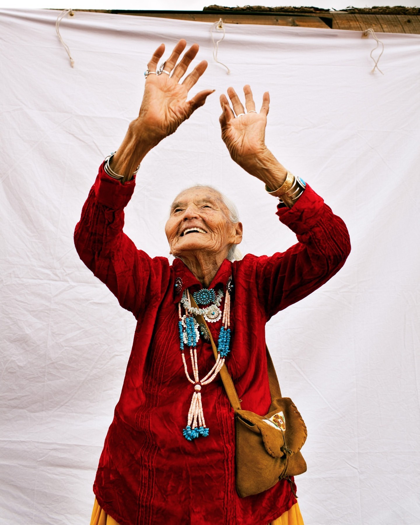 Elderly Indigenous woman in red dress with arms raised, adorned with turquoise jewelry, smiling against a white backdrop.