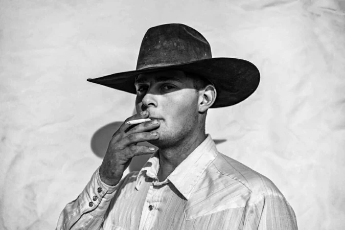 Black and white portrait of a man wearing a cowboy hat and light button-up shirt while smoking a cigarette, shot by Andy Anderson.