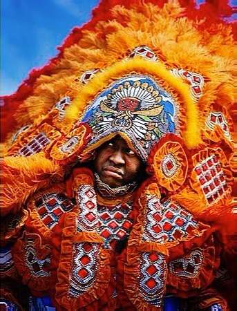 A person in elaborate orange and white feathered costume with beaded headdress. The person appears to be at an outdoor event.
