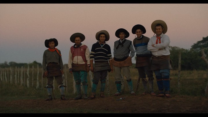Group of six gauchos standing in a rural field at dusk, wearing traditional hats and aprons, with a simple wooden fence and trees in the background.